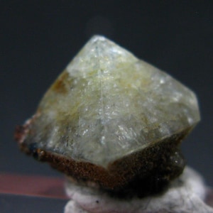 Puede incluir: Un cristal translúcido de color amarillo claro con forma piramidal. El cristal tiene una superficie rugosa y texturizada y una base marrón oscuro. El fondo es un gris oscuro borroso.