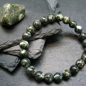Pode incluir: Uma pulseira de contas redondas, salpicadas de verde e branco. Uma pedra semelhante está posicionada ao lado da pulseira. A joia é exibida em uma superfície escura e texturizada.