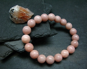 Könnte beinhalten: Ein Armband aus großen, runden, rosa Edelsteinperlen. Das Armband wird auf einer dunklen, strukturierten Oberfläche präsentiert, mit einem Stück rauem, orange-braunem Stein im Hintergrund. Die Perlen haben eine polierte Oberfläche.