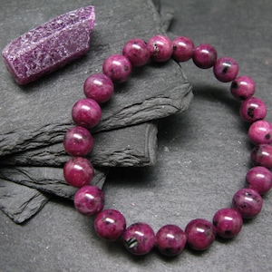 Może przedstawiać: Bransoletka z dużymi, okrągłymi, ciemnoróżowymi koralikami. W pobliżu znajduje się surowy, krystaliczny, fioletowy kamień szlachetny. Bransoletka jest prezentowana na ciemnej, teksturowanej powierzchni, tworząc kontrast z żywymi kolorami biżuterii.