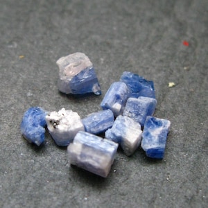 May include: A collection of small, raw kyanite crystals featuring varying shades of blue, ranging from deep indigo to pale sky blue, with some pieces displaying white, opaque mineral inclusions and a translucent, crystalline, and layered texture.