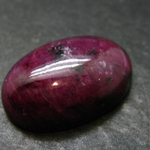 Op de afbeelding: Een ovaalvormige cabochon edelsteen met een diep magenta en donkerpaars kleurenpalet. Het oppervlak is gepolijst en glad, met zichtbare natuurlijke insluitingen, donkere mineraalvlekken en een reflecterende glans over de gebogen bovenkant.