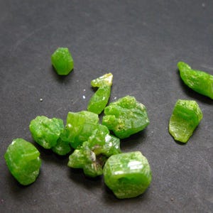 May include: A collection of bright green, crystalline gemstones scattered on a dark surface. The stones vary in size and shape, with a translucent, faceted appearance. The gemstones are likely raw or uncut.
