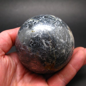 Puede incluir: Una piedra esférica pulida, con aspecto veteado. El orbe presenta una mezcla de patrones gris oscuro, negro y blanco, con finas vetas doradas. La piedra se sostiene en una mano, resaltando su superficie lisa y reflectante. Un artículo decorativo.