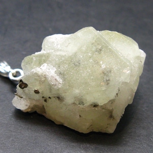 May include: A light green, raw crystal pendant with a silver-colored bail. The crystal has a rough, textured surface with small dark inclusions. The pendant is set against a dark gray background.