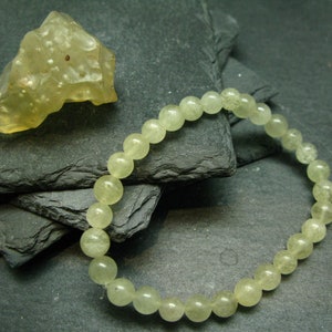 May include: A light green beaded bracelet and a rough-cut gemstone. The bracelet features translucent, round beads. The gemstone is a pale yellow colour with a textured surface. The items are displayed on a dark grey, layered surface.