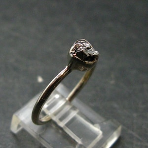 May include: A silver-toned metal ring featuring a single, small, rough-textured stone set in a prong setting. The band is thin and smooth, showcasing a metallic lustre. The ring is positioned at an angle, highlighting the irregular shape of the stone.
