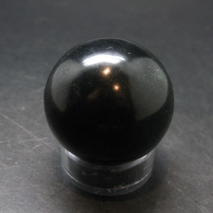 Può includere: Una sfera nera lucida, forse un oggetto decorativo o una pietra preziosa, appoggiata su un supporto trasparente e circolare. La sfera riflette la luce, creando riflessi sulla sua superficie liscia. Lo sfondo è una superficie scura e strutturata.