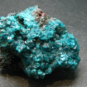 Könnte beinhalten: Ein Cluster aus blaugrünen Kristallen auf einem dunkelgrauen Stein. Die Kristalle sind klein und haben eine raue, strukturierte Oberfläche.