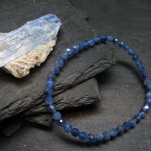 Puede incluir: Una pulsera de cuentas azules con cuentas facetadas se muestra sobre una superficie gris oscuro. Un trozo de cristal de cianita azul en bruto descansa cerca. La pulsera tiene aproximadamente 18 cm de circunferencia.