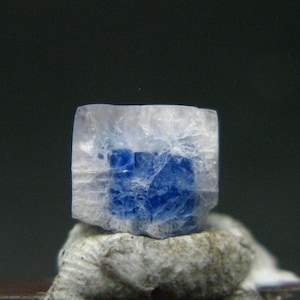 May include: A raw, cubic mineral specimen featuring a translucent, frosted white outer layer that transitions into a vibrant, deep blue crystalline core. The stone displays a distinct geometric shape with visible internal fractures and crystalline textures.