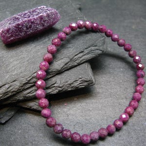 May include: A faceted ruby bracelet and a raw ruby gemstone. The bracelet is made of small, round, deep pink beads. The raw ruby is a rectangular shape with a rough, textured surface. Both are displayed on a dark grey, textured surface.