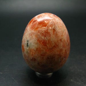 May include: A polished, egg-shaped sunstone mineral specimen featuring a mottled texture with warm orange, reddish-brown, and cream-coloured inclusions throughout its surface, displaying a natural crystalline structure with subtle reflective highlights.