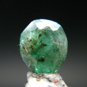 May include: A faceted, translucent green gemstone with visible internal inclusions and dark mineral deposits. The stone features a rounded, irregular shape with multiple polished surfaces that catch the light, displaying varying shades of emerald green.