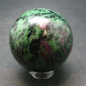 Puede incluir: Una esfera de piedra preciosa verde, negra y rosa. La esfera es lisa y pulida, con un patrón moteado.