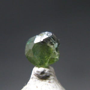 May include: A faceted, green gemstone with a crystalline structure. The gem is a deep, verdant color, with multiple reflective surfaces. It is mounted on a white, textured base, set against a gray background.