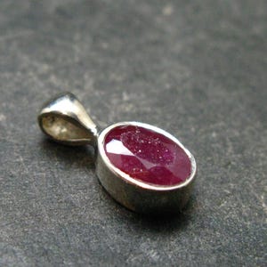 May include: A pendant featuring an oval-shaped, faceted deep red ruby gemstone set in a smooth, polished silver-toned metal bezel. The pendant includes a small, integrated silver-toned metal bail at the top for attachment to a chain or cord.