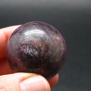 May include: A polished, spherical gemstone with a deep purple hue, accented by streaks of darker shades and hints of gold. The surface reflects light, showcasing the stone's natural patterns and textures. This decorative sphere is a unique addition to any collection.