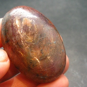 Puede incluir: Una piedra de cobre lisa de forma ovalada con un tono marrón rojizo. La piedra tiene una superficie ligeramente texturizada con remolinos y líneas visibles.