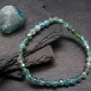 May include: A beaded bracelet with faceted, teal-colored gemstones. A matching, rough-cut gemstone sits nearby on a dark, textured surface. The bracelet is strung on an elastic band, showcasing the individual beads and their varying shades of blue and green.