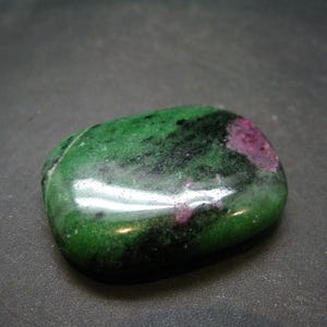 Puede incluir: Una piedra preciosa lisa, verde y negra con acentos rosados. La piedra está pulida y tiene un acabado brillante.