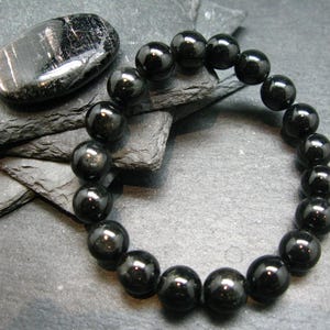 May include: A black beaded bracelet with large, round beads. A polished black stone sits next to the bracelet on a dark gray, textured surface. The bracelet is strung together, forming a circular shape.