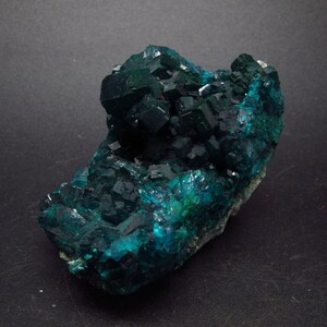 Puede incluir: Un cúmulo de cristales de color verde oscuro, verde azulado y azul con una textura rugosa y natural. Los cristales están dispuestos en un cúmulo, con algunos cristales más grandes que otros. Los cristales tienen un brillo brillante, casi metálico.