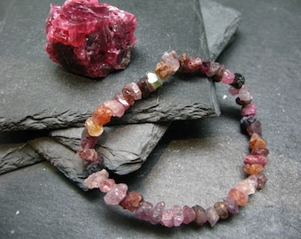 May include: A bracelet made of pink, red, and black gemstone beads. The beads are irregularly shaped and have a rough, natural finish. The bracelet is lying on a dark gray surface.