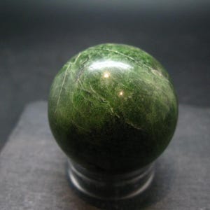 May include: A smooth, green sphere of polished serpentine stone. The stone has a marbled pattern and is displayed on a black stand.