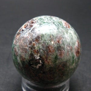 Puede incluir: Una esfera de piedra preciosa pulida con aspecto moteado. La esfera presenta una mezcla de colores verdes, marrones y blancos, creando una estética natural y terrosa. La forma esférica es lisa y reflectante.