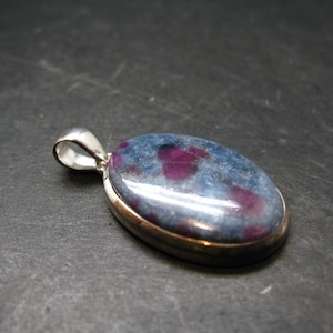 May include: Oval pendant with a silver bail, featuring a blue-grey stone with vibrant red inclusions. The stone is set in a silver-toned metal frame. A piece of jewellery with a natural stone.