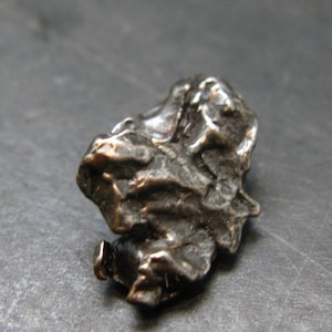 May include: A small, irregularly shaped, metallic object with a rough, textured surface. The object has a dark, bronze-like colour with areas of lighter highlights. The overall form is organic and appears to be a natural specimen, possibly a mineral or a small sculpture.