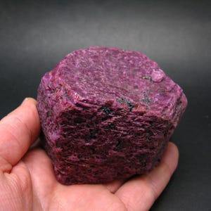 Könnte beinhalten: Ein roher, ungeschliffener Rubin-Edelstein mit tiefroter bis violetter Farbe. Der Naturstein hat eine strukturierte Oberfläche und eine unregelmäßige Form mit kleinen schwarzen Einschlüssen. Der Rubin wird in einer Hand vor dunklem Hintergrund gehalten.