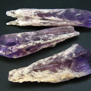 Puede incluir: Tres puntas de cristal de amatista con un patrón de color púrpura y blanco. Los cristales son en bruto y tienen una textura natural y rugosa.