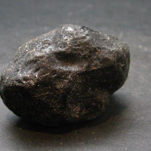 Puede incluir: Una roca oscura de forma irregular con una superficie rugosa y texturizada. La roca es de color negro intenso y parece ser una piedra natural. La imagen es un primer plano, que destaca los detalles de la superficie de la roca.