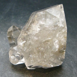 Peut inclure: Un amas de cristaux de quartz transparents, formé naturellement, présentant une pointe de terminaison proéminente à facettes multiples et des cristaux secondaires plus petits sur le côté, avec des fractures internes et des surfaces réfléchissantes.