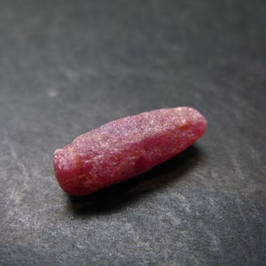 May include: A rough, elongated ruby gemstone, displaying a deep reddish-pink hue. The natural, unpolished stone has a slightly irregular shape and a textured surface. The ruby is set against a dark background.