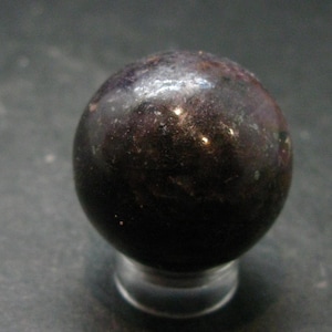 Può includere: Una pietra sferica lucida con una tavolozza di colori viola scuro e nero brunastro. La superficie presenta sottili macchie metalliche riflettenti e inclusioni minerali naturali in tutta la sfera, poggiata su un piccolo supporto circolare trasparente.