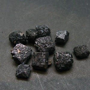 May include: A group of black, faceted, rough, natural, gemstone crystals. The crystals are various sizes and shapes.