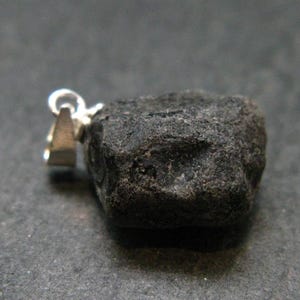 Puede incluir: Un colgante de piedra en bruto, de color gris oscuro y textura rugosa, con una forma irregular y dentada. La piedra está sujeta a una anilla de metal plateado pulido, que proporciona un orificio para colgar la pieza en una cadena.