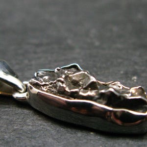 May include: A silver pendant featuring a rough, dark, textured metallic stone set within a polished, smooth silver frame. The pendant includes a silver bail at one end for attachment, showcasing a contrast between the raw, uneven surface of the stone and the reflective, shiny metal border.