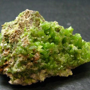 Puede incluir: Un espécimen mineral en bruto que presenta un grupo de estructuras cristalinas de color verde lima vibrante que crecen a partir de una matriz rocosa texturizada de tonos tierra. La parte cristalina muestra un aspecto facetado y translúcido.