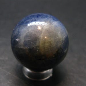 Peut inclure: Une pierre précieuse sphérique polie, présentant un dégradé de couleurs allant du bleu foncé en haut à une teinte plus foncée à la base. La sphère est montée sur un petit support argenté, mettant en valeur sa surface lisse et réfléchissante.