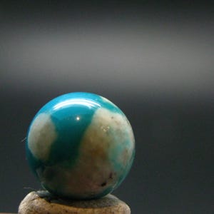 Può includere: Una pietra preziosa sferica lucidata caratterizzata da un vibrante colore blu turchese con inclusioni irregolari di colore bianco nuvoloso e marrone chiaro su tutta la superficie liscia e lucida, che mostra un motivo naturale con una finitura riflettente.