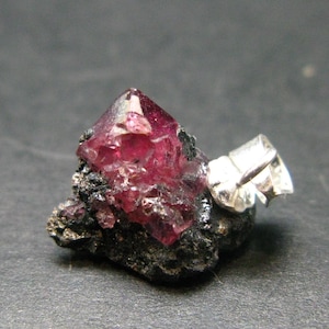 Puede incluir: Primer plano de un espécimen mineral natural con un cristal rosa intenso y facetado. El cristal está sobre una matriz oscura y rugosa con un accesorio plateado, mostrando su belleza natural y formación geológica.