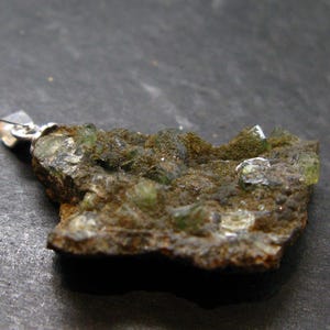 May include: A natural, rough-cut gemstone pendant. The pendant features a cluster of green crystals embedded in a matrix of brown and gray rock. A silver-colored bail is attached for hanging.