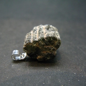 May include: A dark green, rough, and textured stone pendant with a silver bail. The stone has a striated pattern and is attached to a silver bail.