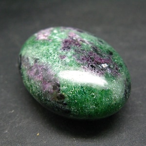 May include: A green and purple gemstone with a smooth, polished surface. The stone has a speckled appearance with dark green, light green, purple, and black flecks.