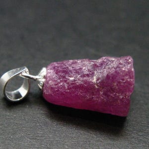 May include: A magenta-coloured, uncut ruby pendant with a silver bail. The ruby has a rough, textured surface. The pendant is displayed against a dark background.