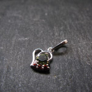 May include: A silver pendant with a heart-shaped design. The pendant features a green gemstone at the center, surrounded by smaller red gemstones. The pendant is suspended from a silver bail. The background is a dark, textured surface.